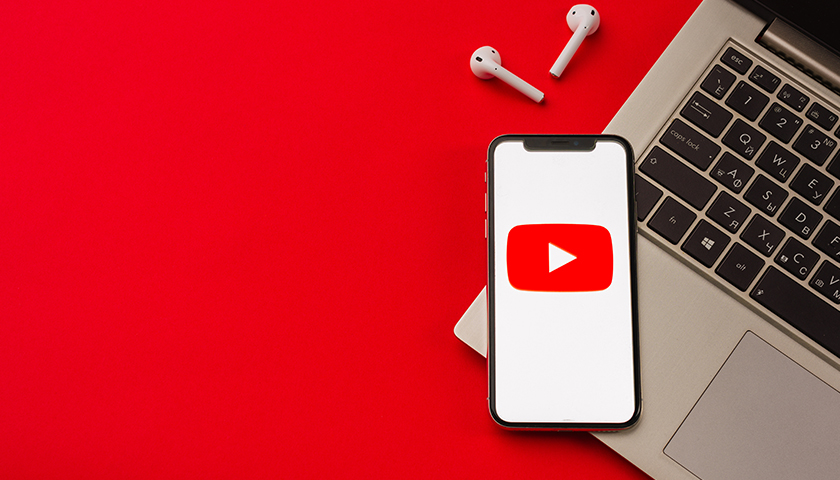 Smartphone displaying the YouTube logo, resting on a laptop with wireless earbuds on a red background.